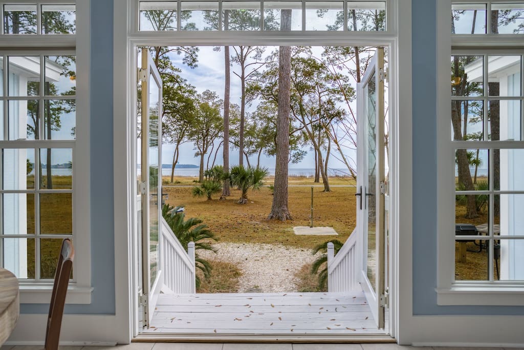 Open French doors of a light coastal cottage opening to a white porch and gravel path flanked by palmettos and pine trees, leading to a grassy salt marsh and calm bay waterfront view.