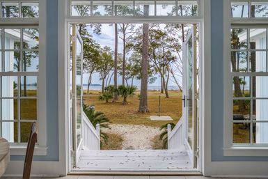 Open French doors of a light coastal cottage opening to a white porch and gravel path flanked by palmettos and pine trees, leading to a grassy salt marsh and calm bay waterfront view.