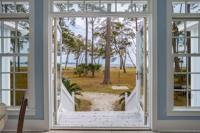 Open French doors of a light coastal cottage opening to a white porch and gravel path flanked by palmettos and pine trees, leading to a grassy salt marsh and calm bay waterfront view.
