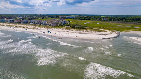 Aerial view of a coastal sandy beach with rolling ocean waves, beachgoers and umbrellas, a rocky groin, a row of oceanfront homes and green marshland under a partly cloudy sky