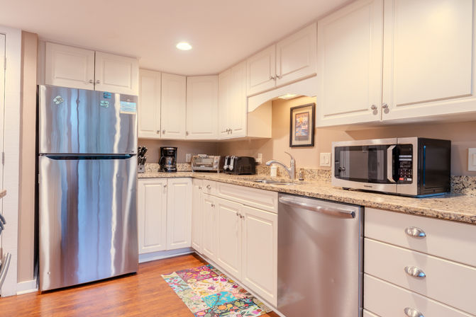 Bright modern L-shaped kitchen with white shaker cabinets, granite countertops, stainless steel refrigerator and dishwasher, microwave, toaster and coffee maker on counter, hardwood floor and colorful runner rug.