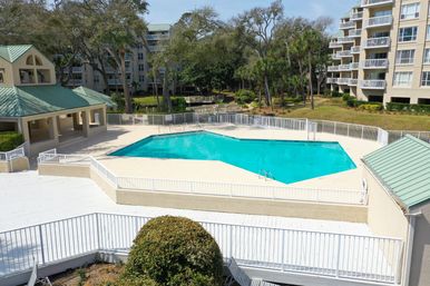Sunny outdoor turquoise pool at a coastal condominium complex with white deck railings, green-roof pavilions, balconies and palm trees.