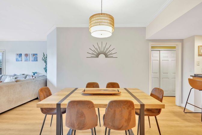 Open-concept modern dining room in a contemporary home with a rectangular wood table, six rust-brown chairs, woven pendant light, metal sunburst wall art, light wood floors and a peek into the neutral-toned living area.