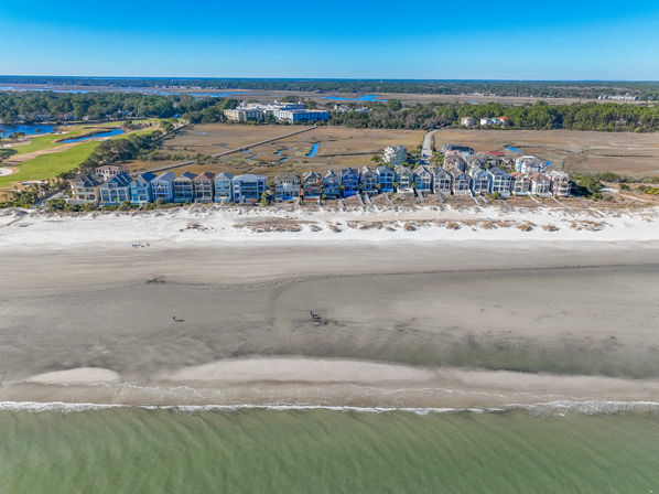 Aerial view of a sunny row of colorful oceanfront homes along a wide sandy beach, gentle green waves in the foreground and marshland and golf course visible behind under a clear blue sky