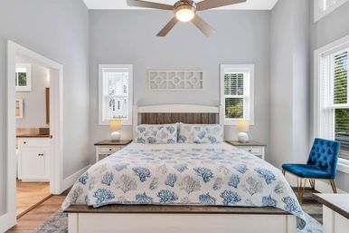 Sunlit modern coastal bedroom with white wood bed, blue coral-patterned quilt and pillows, matching nightstands with lamps, ceiling fan, and a tufted blue accent chair by the window.