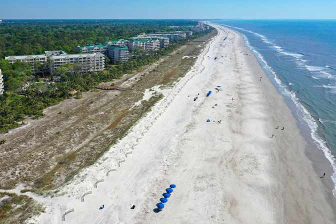 Aerial view of a long sandy beach with ocean waves, dune vegetation and beachfront condos; scattered blue umbrellas and small groups of beachgoers along the shoreline.