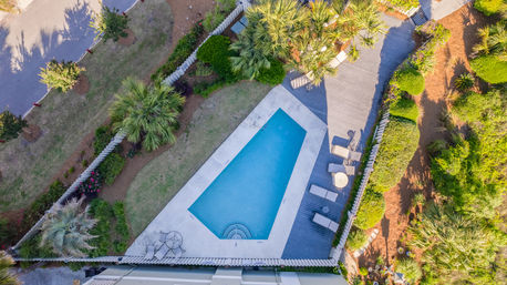 Aerial view of a geometric outdoor swimming pool with a concrete border and wooden deck, sun loungers and table, framed by palm trees and lush landscaped yard.