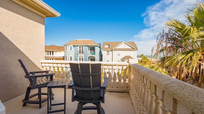 Sunny ocean-view balcony with two wooden lounge chairs, decorative balustrade, palm tree and pastel coastal houses beyond.