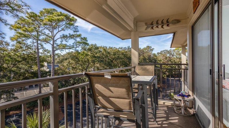Sunny balcony patio with high-top table and two chairs, sliding glass door to condo, overlooking pine-tree-lined neighborhood under a blue sky.