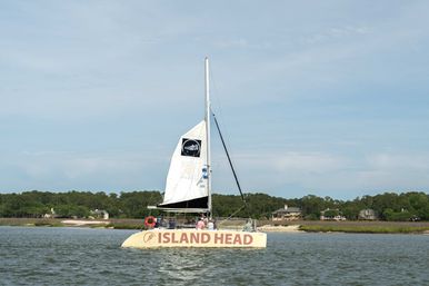 Yellow catamaran sailboat with tall white sail and passengers cruising in a calm coastal inlet beside marshy shoreline and waterfront homes under a blue sky.
