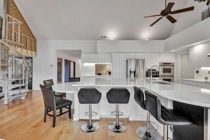 Bright open-concept modern kitchen with large white quartz island, five black leather bar stools, stainless steel appliances and double oven, hardwood floors, spiral staircase and ceiling fan.