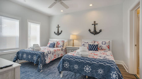 Sunlit coastal bedroom with two twin beds in navy-and-white nautical quilts, anchor wall decor above a white nightstand with lamp, folded towels on the beds, ceiling fan and bright windows for a beach vacation rental vibe.