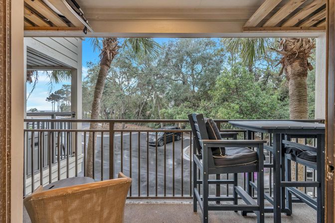 Coastal balcony with wicker chair and cushioned high-top bar table and chairs, metal railing overlooking palm trees, parking area, and blue sky.