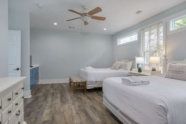 Spacious coastal-style vacation bedroom with two queen beds in white linens, wood-look floors, ceiling fan, bedside lamps, bench and sunlit windows with blinds