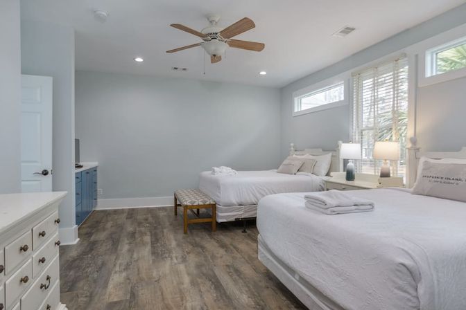 Spacious coastal-style vacation bedroom with two queen beds in white linens, wood-look floors, ceiling fan, bedside lamps, bench and sunlit windows with blinds