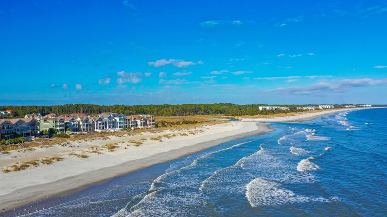 Aerial view of beachfront homes lining a wide sandy beach and dunes with gentle ocean waves rolling under a bright blue sky