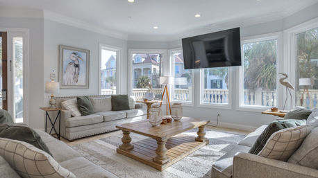 Bright coastal living room with bay windows overlooking palm trees and seaside homes, two light-gray sofas with throw pillows, rustic wooden coffee table, patterned rug, floor lamps and a wall-mounted TV for a relaxed beachy vibe.