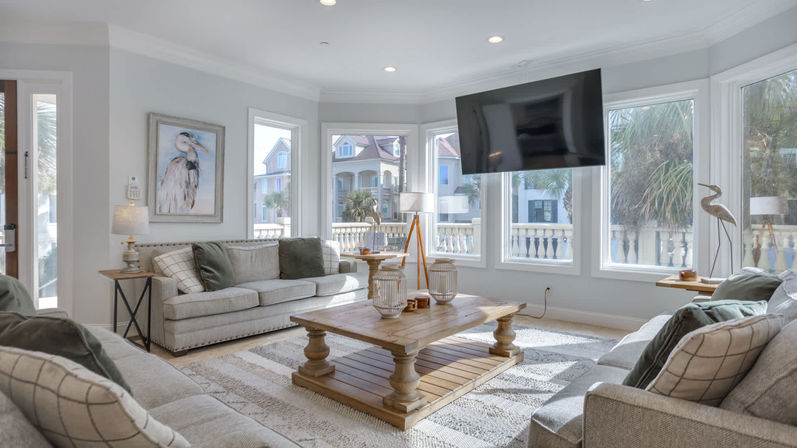 Bright coastal living room with bay windows overlooking palm trees and seaside homes, two light-gray sofas with throw pillows, rustic wooden coffee table, patterned rug, floor lamps and a wall-mounted TV for a relaxed beachy vibe.
