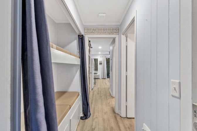 Coastal beach-cottage hallway with built-in bunk beds curtained in navy, light wood-look flooring, pale shiplap walls and a 'Beach Rules' sign leading to a bright living area.