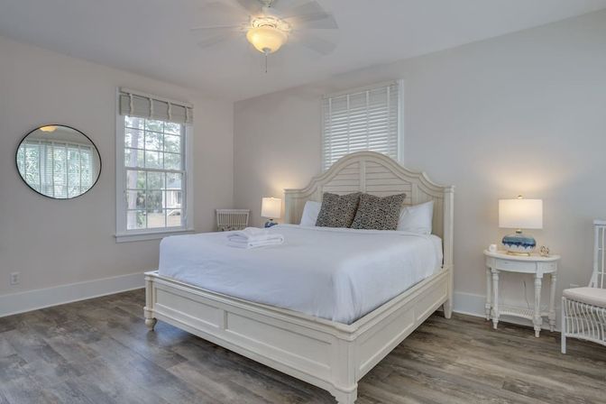 Sunlit coastal-style guest bedroom with white wooden bed and headboard, crisp white bedding topped with folded towels and patterned pillows, wood-look floors, round wall mirror, two windows and bedside tables with lamps.