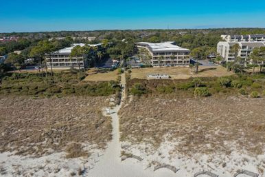 Aerial view of a sandy beach access path through dunes and coastal grasses leading to beachfront low-rise condos and pine trees under a clear blue sky.