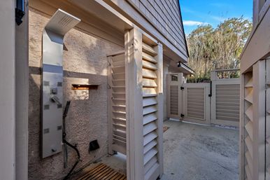 Sunlit private outdoor shower with a modern stainless steel rainfall shower panel, wooden slatted mat and beige louvered privacy panels opening to a fenced courtyard under a blue sky