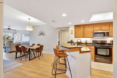 Bright open-concept kitchen and dining area with wood floors, granite breakfast bar and leather stools, warm wood cabinets, stainless appliances, dining table and balcony with palm trees