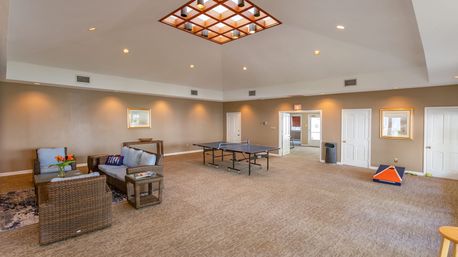 Spacious indoor community game room with ping-pong table at center, wicker seating with blue cushions, cornhole board, skylight grid, recessed lighting, beige walls and carpet — bright clubhouse recreation area.