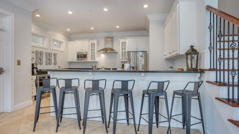 Modern open-concept kitchen with white cabinets, stainless steel appliances, black granite breakfast bar lined with six gray metal bar stools, tiled floor and adjacent staircase