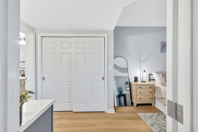 Bright modern bedroom interior with white sliding closet doors, light wood floors, neutral gray walls, a leaning full-length mirror and a wooden nightstand with lamp