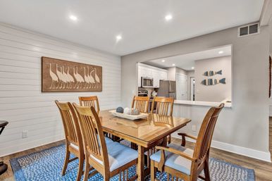 Coastal-style dining room with wooden six-seat table and blue-upholstered chairs on a patterned blue rug, shiplap accent wall featuring bird art, and an open passthrough to a white kitchen with stainless appliances.
