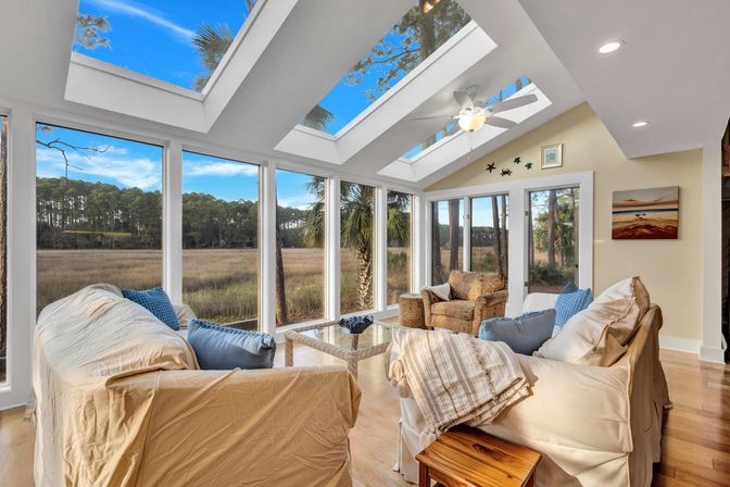 Sun-drenched coastal sunroom with skylights and floor-to-ceiling windows overlooking marshland and pine trees, cozy sofas, armchair and ceiling fan.