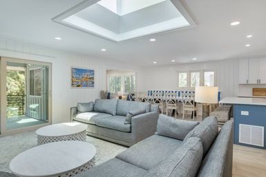 Bright coastal-style open-plan living room with skylight, gray sofas, round white coffee tables, dining area, and balcony view.