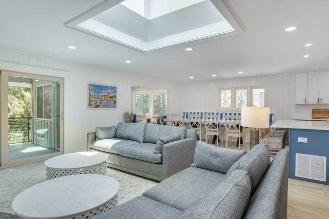 Bright coastal-style open-plan living room with skylight, gray sofas, round white coffee tables, dining area, and balcony view.