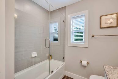 Modern residential bathroom with white tub and frameless glass shower door, gray horizontal tile surround, small window with white blinds, brass fixtures and wood-look flooring