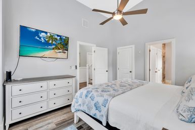 Bright modern coastal master bedroom with light gray walls, ceiling fan, wall-mounted TV showing a tropical beach, white dresser, king bed with blue coral-patterned bedding and wood-look floors