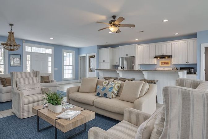 Bright coastal-style open-concept living room and kitchen with light blue walls, beige sofa, striped armchairs, wooden coffee table, blue rug, white shaker cabinets and stainless steel appliances.
