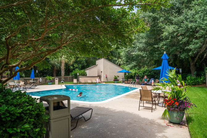 Sunny outdoor community swimming pool with turquoise water, lounge chairs and blue umbrellas on a landscaped deck, large shade trees and tropical plants, swimmer in the pool and people relaxing near a beige pool house.
