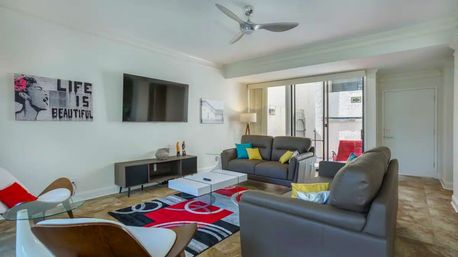 Sunny contemporary condo living room with gray leather sofas, colorful throw pillows, wall-mounted flat-screen TV, glass coffee table, red-and-black abstract rug, ceiling fan and sliding glass balcony doors.
