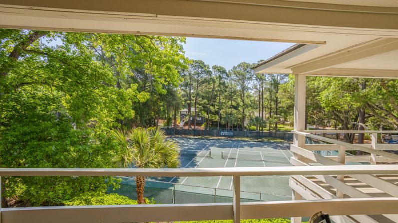 Sunny balcony view overlooking fenced community tennis courts framed by white rails, palm and pine trees.