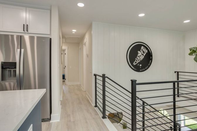 Bright modern coastal-style hallway and kitchen area with light wood floors, stainless steel refrigerator and white quartz countertop on the left, white shiplap wall with round black decorative sign, and black metal railing overlooking a staircase.