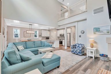Bright coastal-style open-concept living room and kitchen with vaulted two-story ceiling, large turquoise sectional sofa, patterned armchair, white kitchen island with barstools, dining area and wood-look floors.
