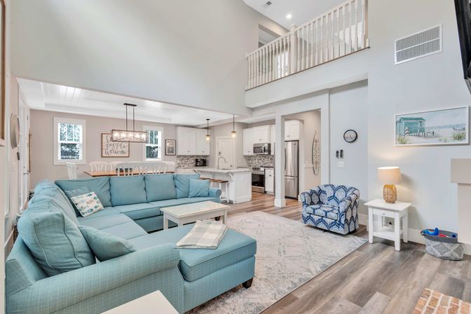 Bright coastal-style open-concept living room and kitchen with vaulted two-story ceiling, large turquoise sectional sofa, patterned armchair, white kitchen island with barstools, dining area and wood-look floors.