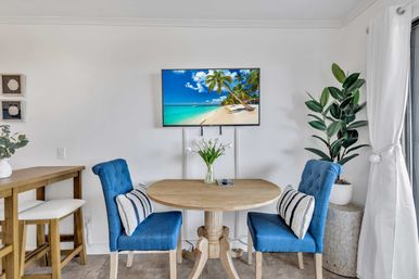 Bright coastal dining nook with round wooden table, two blue upholstered chairs with striped pillows, wall-mounted TV showing a tropical beach, tall potted plant and nearby bar stools.