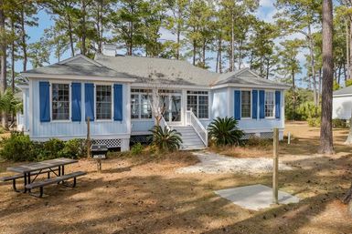 Light blue coastal cottage with bright blue shutters and white steps, nestled among tall pine trees with a picnic table, charcoal grill, sandy yard and palm-like shrubs — charming beachside bungalow vibe.
