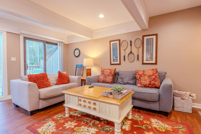 Cozy, sunlit living room with hardwood floors, beige walls, gray and cream sofas with red patterned pillows, cream wood coffee table on a floral red rug, vintage tennis rackets on the wall and a sliding glass patio door.