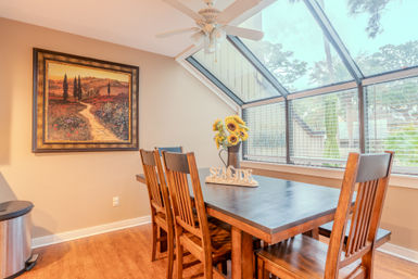 Cozy sunlit dining nook with wooden table and chairs beneath slanted glass skylight, sunflower bouquet and “SEASIDE” sign on the table, framed landscape painting on the wall.