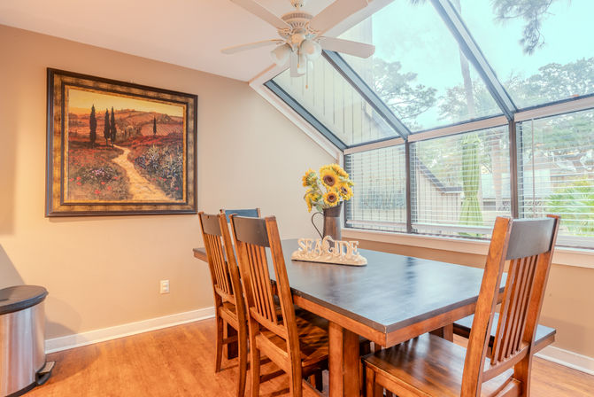 Cozy sunlit dining nook with wooden table and chairs beneath slanted glass skylight, sunflower bouquet and “SEASIDE” sign on the table, framed landscape painting on the wall.