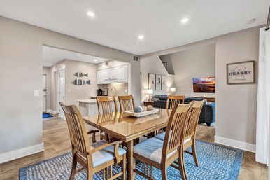 Bright open-concept coastal-style dining room with wooden six-seat table on a blue rug, beachy fish wall decor and "No Crabby" sign, white kitchen cabinets and breakfast bar, adjacent living area with gray sofas, hardwood floors and recessed lighting.