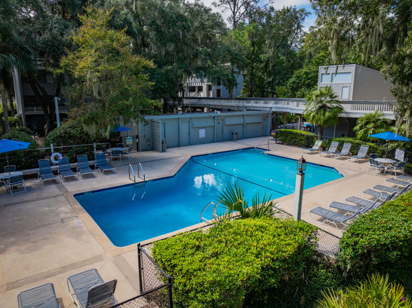 Outdoor rectangular community swimming pool with sparkling blue water, rows of sun loungers and blue umbrellas, palm trees and leafy walkways in a residential courtyard.